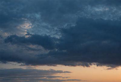 The primary subject of the image is a cloudy sky with dark clouds. The setting is a sunset, creating a dramatic backdrop. The visual style is a photo, capturing the beauty of the cloudy sky during the sunset. The colors and mood of the image are dominated by the dark clouds and the warm hues of the sunset, creating a moody and dramatic atmosphere.