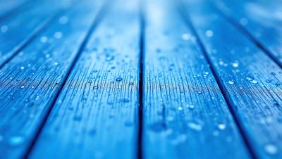The primary subject of the image is a wooden deck with a blue surface. The setting is an outdoor environment, possibly a beach or a patio. The visual style is a photo, capturing the raindrops on the wooden surface. The dominant colors are blue and white, creating a serene and calming atmosphere.