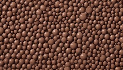 The primary subject of the image is a close-up of a chocolate-covered candy, which appears to be a chocolate-covered peanut. The setting is a brown surface, possibly a wall or a table, with a pattern of chocolate balls or marshmallows. The visual style is a close-up, and the colors are predominantly brown, giving the image a warm and inviting atmosphere.