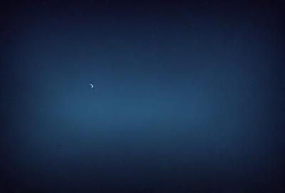 The primary subject of the image is a moon, which is visible in the sky. The setting is a dark, cloudy night with a blue sky. The visual style is a photo, capturing the moon and the night sky. The colors and mood of the image are predominantly dark and moody, with the blue sky providing a contrasting element.