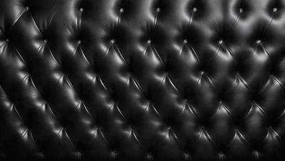 The primary subject of the image is a black leather couch. The setting is a room with a black background, which creates a contrast with the couch. The visual style is a close-up of the couch, showcasing its texture and design. The colors and mood of the image are predominantly black, giving it a sleek and modern appearance.