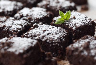 The image features a close-up of a delicious chocolate cake with white frosting. The cake is topped with a single mint leaf, adding a touch of greenery to the dessert. The cake is placed on a dining table, which occupies the majority of the background. The visual style of the image is a close-up, emphasizing the details of the cake and the mint leaf. The colors and mood of the image are warm and inviting, with the rich chocolate and white frosting creating a mouth-watering appearance.