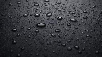 The primary subject of the image is a close-up of a black surface with numerous droplets of water on it. The setting is a dark background, and the visual style appears to be a photo. The colors and mood of the image are predominantly dark, with the droplets of water being the main focus.