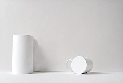 The primary subject of the image is a white coffee cup. The cup is placed on a white surface, which could be a table or a wall. The setting appears to be a simple and clean environment, with no additional objects or distractions. The visual style of the image is a close-up of the coffee cup, emphasizing its shape and texture. The colors in the image are predominantly white, giving the scene a clean and minimalist appearance.