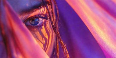 The primary subject of the image is a woman with long, curly hair. She is wearing a purple shawl and has a blue eye. The setting is a room with a pink background. The visual style is a close-up of the woman's face, and the colors and mood are warm and inviting.