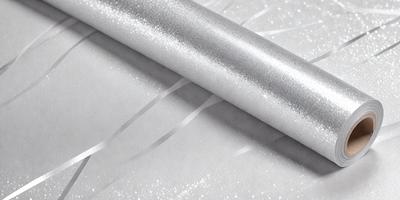 The primary subject of the image is a roll of silver glitter paper. The paper is shiny and metallic, giving it a reflective and glittery appearance. The setting is a table, and the visual style is a close-up of the paper roll. The colors and mood of the image are predominantly silver and reflective, creating a sense of shine and glamour.
