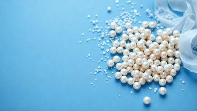 The primary subject of the image is a pile of white pearls. The pearls are sitting on a blue surface, which serves as the setting or background. The visual style of the image is a close-up of the pearls, showcasing their details and texture. The colors in the image are predominantly white and blue, creating a clean and elegant atmosphere.
