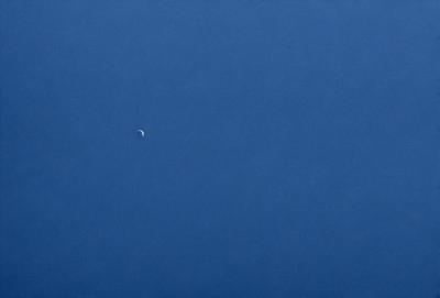 The primary subject of the image is the moon, which is visible in the sky. The setting is a vast, open space with a clear blue sky. The visual style is a photo, capturing the moon in its natural environment. The colors and mood of the image are predominantly blue, creating a serene and peaceful atmosphere.