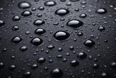 The primary subject of the image is a close-up of a rainy day, with a focus on the raindrops on a black surface. The setting is a rainy day, and the visual style is a close-up of the raindrops. The colors and mood of the image are predominantly black and white, with the raindrops creating a sense of depth and texture.