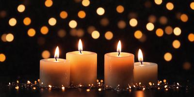 The primary subject of the image is a group of three lit candles placed on a table. The setting is a dark room with a patterned background, creating a cozy and intimate atmosphere. The visual style is a close-up of the candles, emphasizing their glow and the details of their flames. The colors and mood of the image are predominantly warm and inviting, with the candles providing a soft and flickering light source.