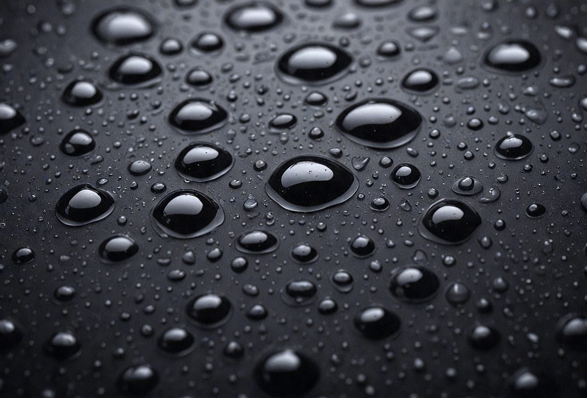 The primary subject of the image is a close-up of a rainy day, with a focus on the raindrops on a black surface. The setting is a rainy day, and the visual style is a close-up of the raindrops. The colors and mood of the image are predominantly black and white, with the raindrops creating a sense of depth and texture.