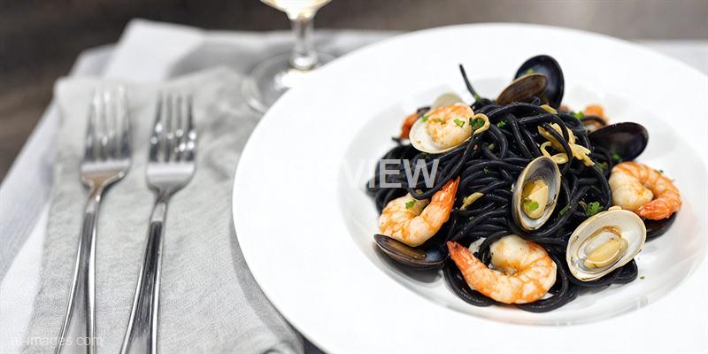 The image shows a white plate filled with black pasta (likely squid ink pasta) topped with shrimp, clams, and garnished with small green herbs. The plate is placed on a white cloth napkin, with two forks to the left and a wine glass in the background, suggesting a fine dining setting.